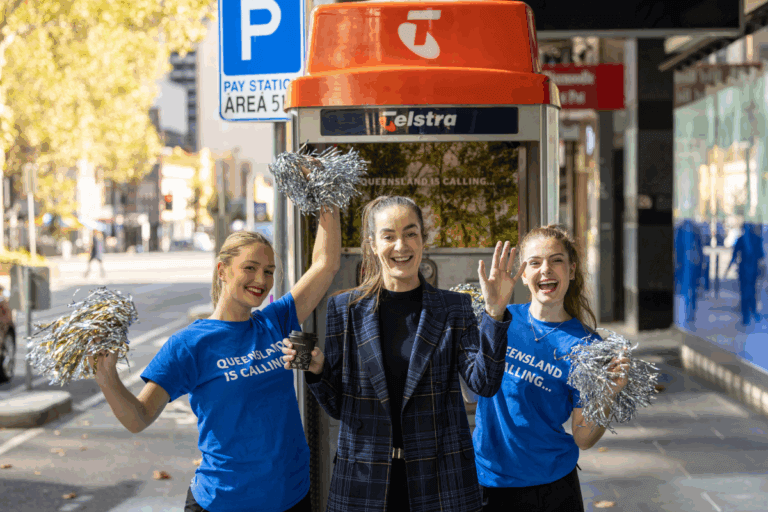 Three women, including two Queensland cheerleaders holding silver pom-poms, smiling in front of a Queensland-branded Telstra phone booth in Melbourne CBD.