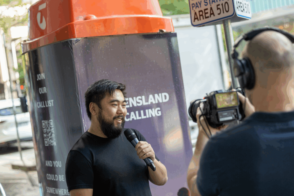 A man speaking into a microphone while being filmed in front of a Queensland-branded Telstra phone booth.