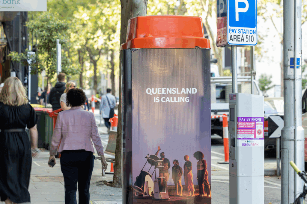 Pedestrians walk past a Queensland-branded Telstra phone booth reading “Queensland is Calling” on a busy Melbourne city street.
