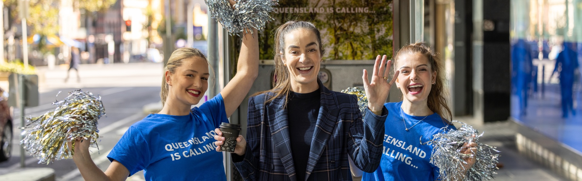 Three women, including two Queensland cheerleaders holding silver pom-poms, smiling in front of a Queensland-branded Telstra phone booth in Melbourne CBD.