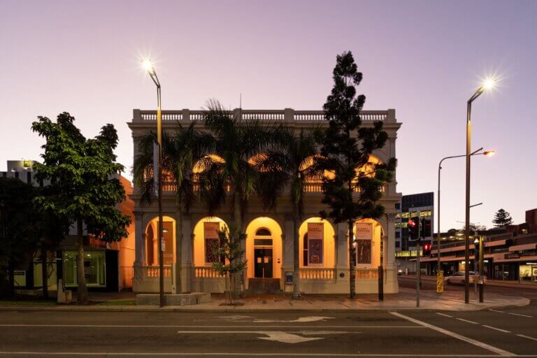 Art gallery at sunset with a purple sky. Archways lit with yellow downlights while trees frame the windows.