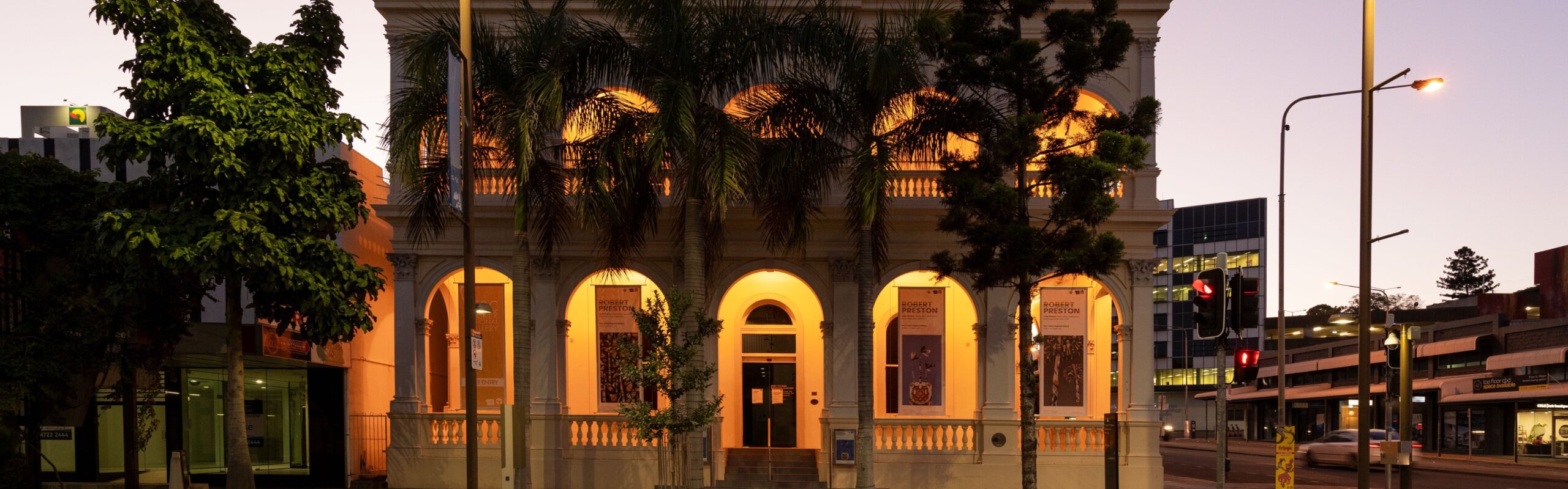 Art gallery at sunset with a purple sky. Archways lit with yellow downlights while trees frame the windows.