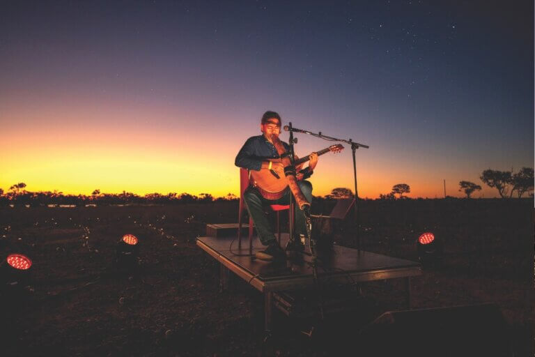 A gradient sunset sky with a musician sitting at the centre playing a didjeridoo and guitar.