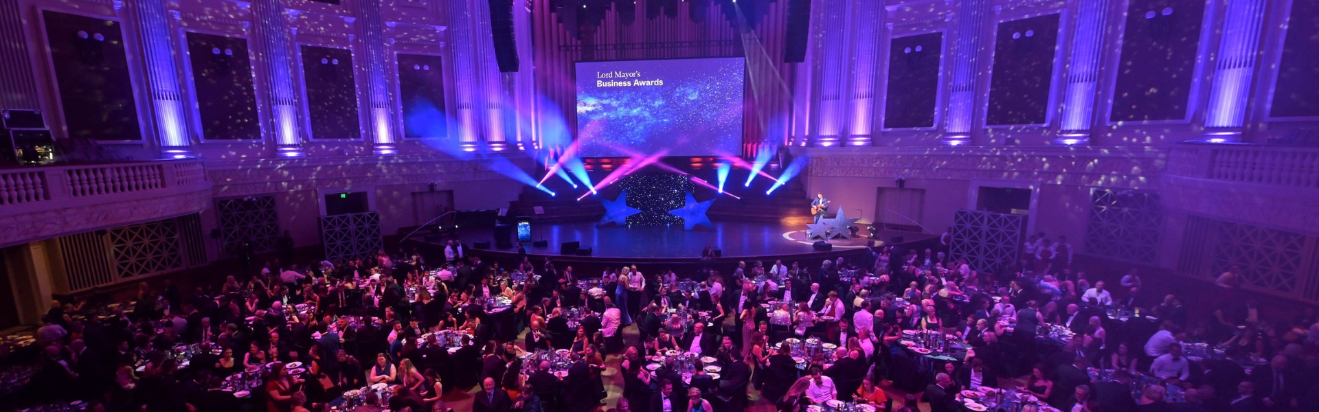 2025 Lord Mayor’s Business Awards Finalists Announced Brisbane City Hall at 2024 Lord Mayor's Business Awards. Stage illuminated with blue, pink and purple lights. Audience with guests seated at round tables.