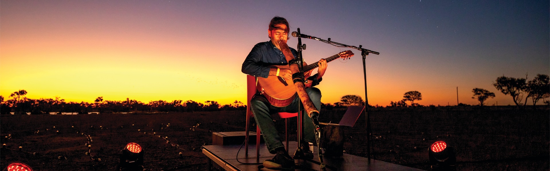 A gradient sunset sky with a musician sitting at the centre playing a didjeridoo and guitar.