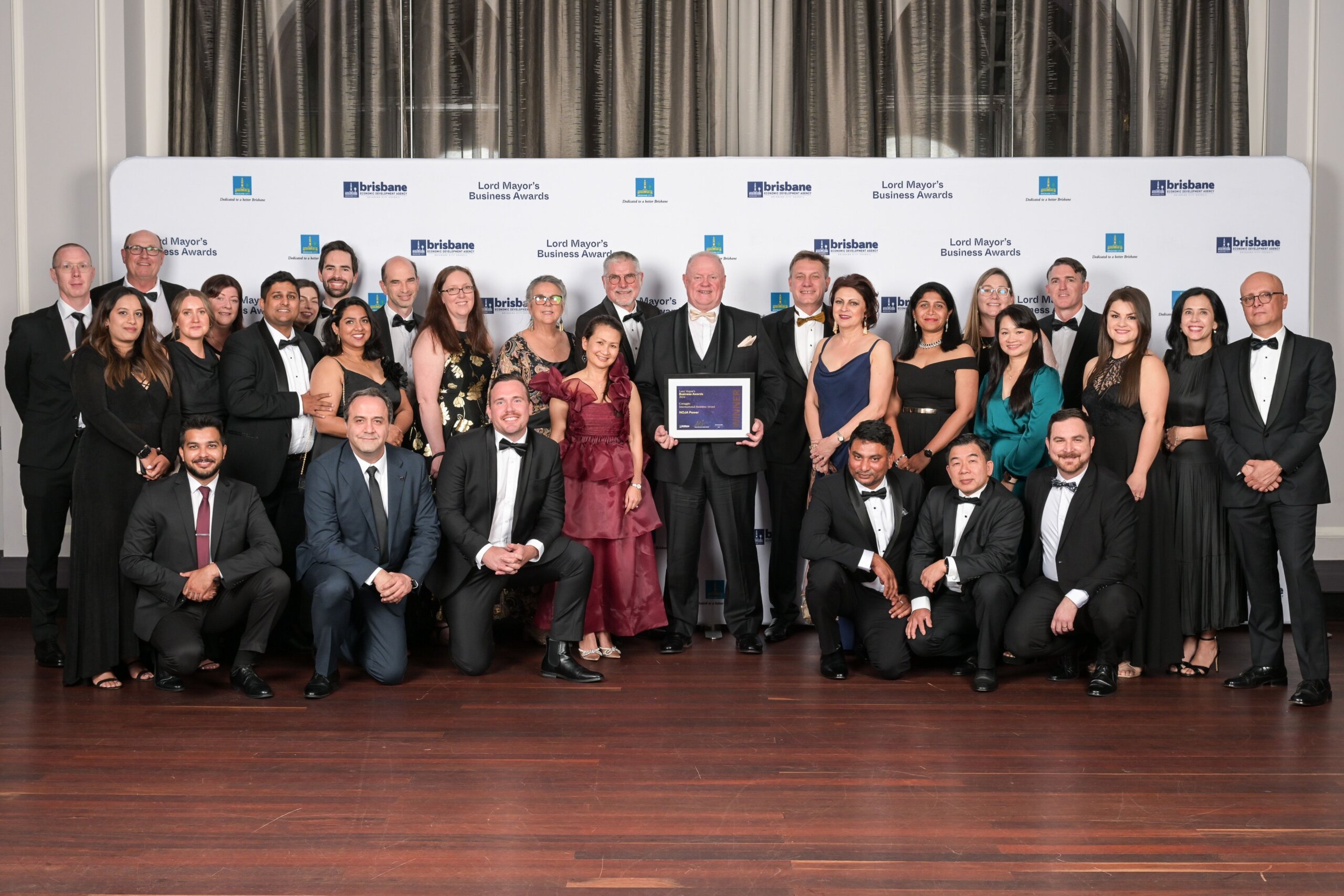 LMBA 2024 NOJA Power 2024 Coraggio International Business Award Winner NOJA Power team pictured ahead of LMBA banner. People wearing formal attire and smiling. Placard held by a man standing in the middle of the team.