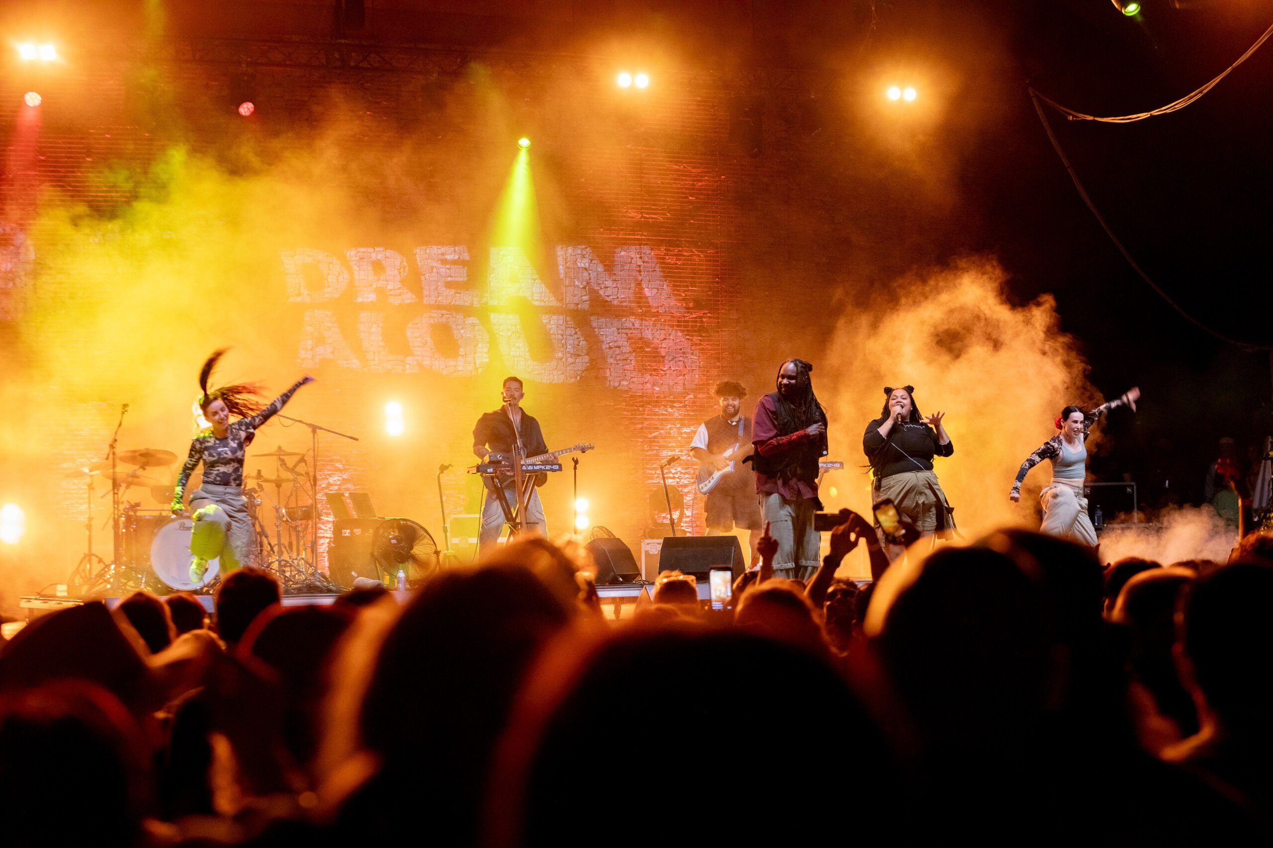 A vibrant stage with yellow lighting, four performers singing and gesturing to crowd.