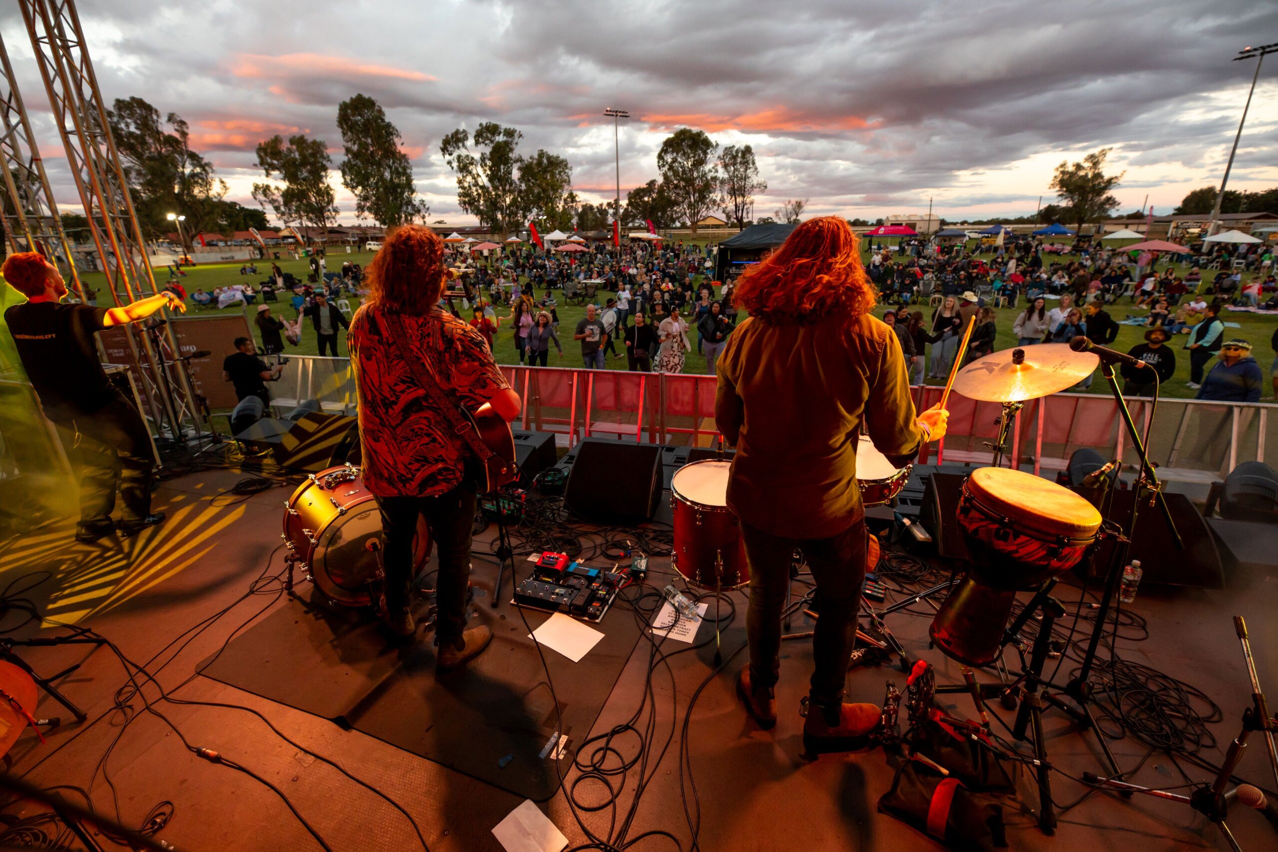 A stage with musicians facing towards an audience. Sunset casting pink light on grey clouds, audience standing on grass and musicians lit by orange stage light.
