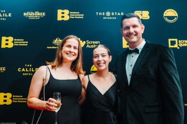 Three people smiling in front of a navy blue media wall at the Brisbane International Film Festival. They are all wearing black and looking towards the camera.
