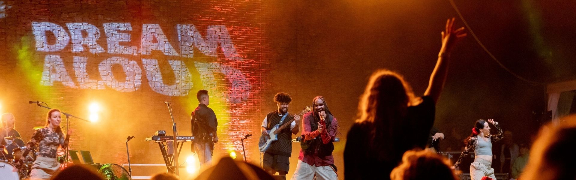 Queensland Music Trails The Tropics. Stage illuminated by orange lights with 'Dream Aloud' written on it. Performers lit by stage lights look towards the audience. One audience member has arm raised towards stage.