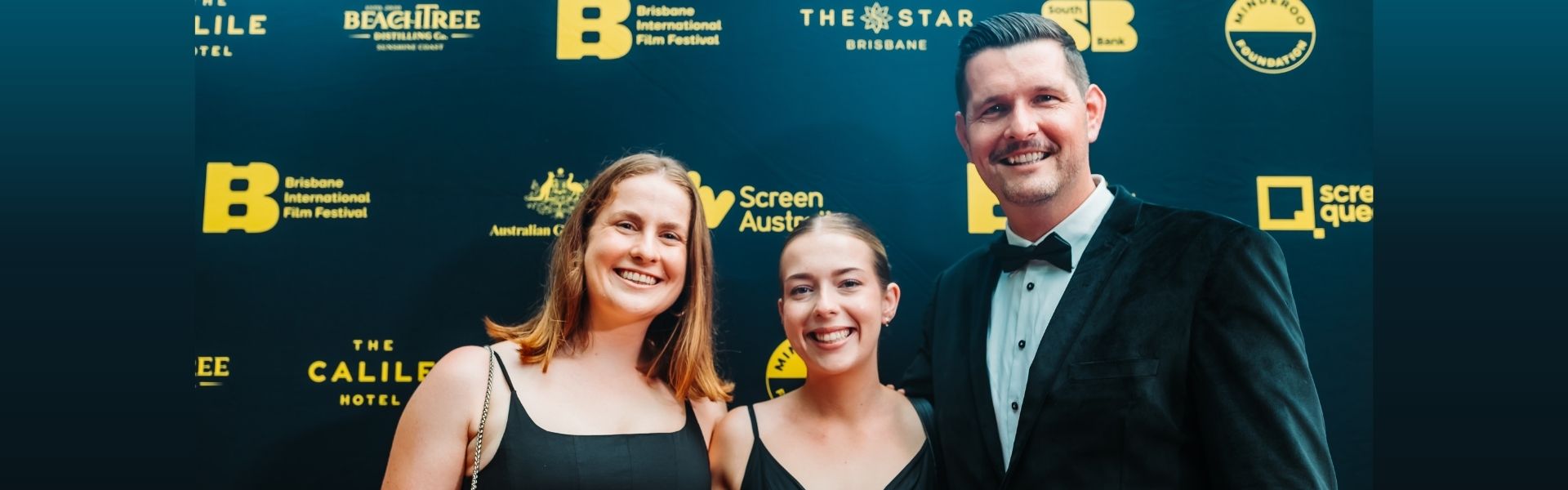Three people smiling in front of a navy blue media wall at the Brisbane International Film Festival. They are all wearing black and looking towards the camera.