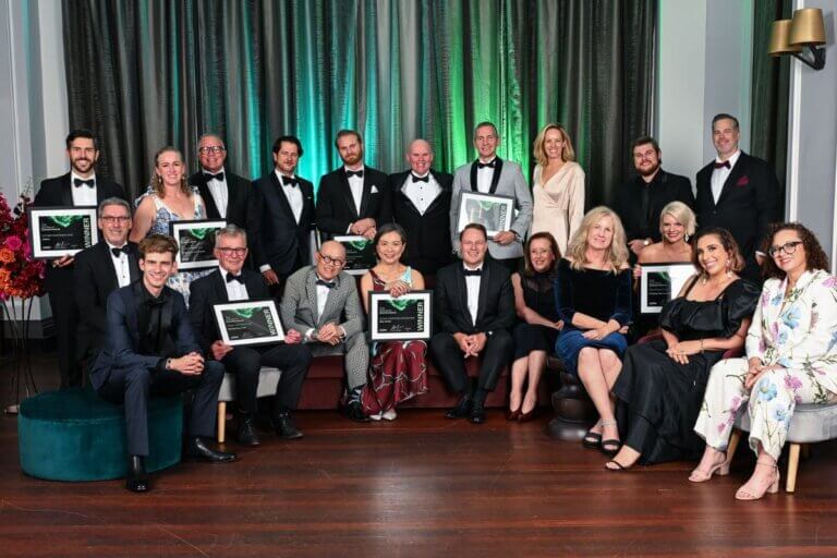 Image of the 2025 Lord Mayor's Business Awards winners. 21 people pictured holding their framed awards. Green light illuminating background.