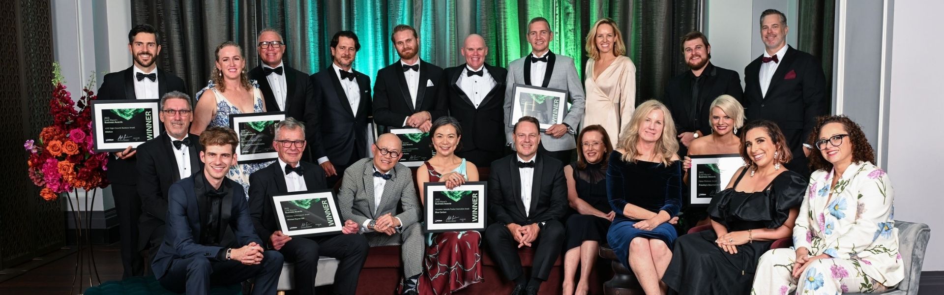 Image of the 2025 Lord Mayor's Business Awards winners. 21 people pictured holding their framed awards. Green light illuminating background.