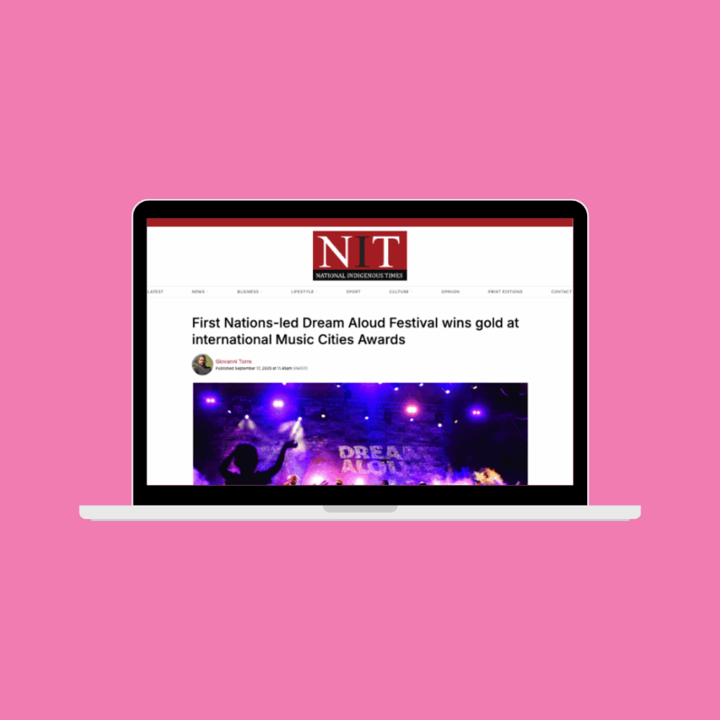 Pink background with image of a laptop within. Laptop screen has an article from National Indigenous Times about Queensland Music Trails The Tropics with title 'First Nations-Led Dream Aloud Festival Wins Gold at International Music Cities Awards.'