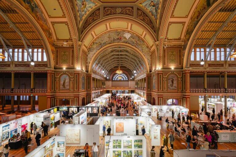 Affordable Art Fair Melbourne. Wide image inside Melbourne's Royal Exhibition Building with white stalls full of art on ground level.